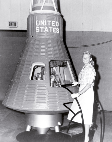 Jerrie Cobb pilotua Mercury proiektuko espazio-kapsularen alboan. Cobbek astronauta izateko proba fisiko eta mediko guztiak gainditu zituen, FLAT programako beste emakume batzuek bezala. Baina ez zieten espaziora joateko aukerarik eman. (Arg.: NASA)