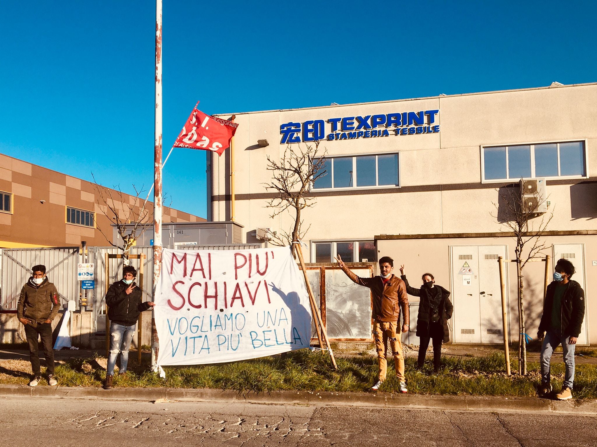 Texprint fabrikaren atarian grebari hilabete luzez eutsi dioten langile etorkinak.  “Esklabo sekula gehiago ez. Bizitza ederragoa nahi dugu” dio banderolak.
argazkia: Produzioni dal Basso